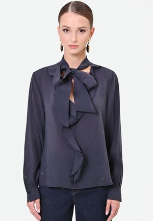 Donna con capelli pettinati all'indietro indossa una camicetta blu navy scuro con una grande fiocco al collo e maniche lunghe, abbinata a jeans scuri.