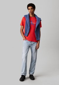 Camiseta de algodón roja con logo azul, llevada con un sudadera azul sobre los hombros. Jeans de mezclilla azul claro y zapatos negros.