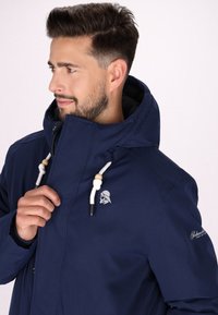 Giacca blu navy con cappuccio, chiusure con cordino e un logo sul petto. Presenta una texture liscia e un design aderente.
