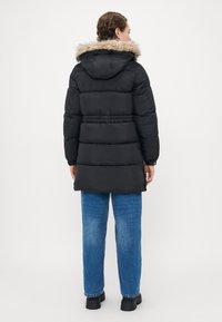 Donna che indossa un cappotto imbottito nero con cappuccio foderato in pelliccia, jeans blu e stivali neri, in piedi di spalle contro uno sfondo bianco semplice.
