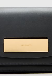 Borsa a mano in pelle nera con placca in metallo dorato. Presenta una texture liscia e un design minimalista con chiusura a patta e branding in rilievo.