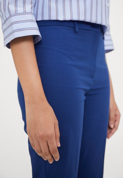 Un pantalon bleu sur mesure présente une texture lisse, une coupe slim, des poches avant et une fermeture éclair, associé à une chemise à manches à rayures claires.