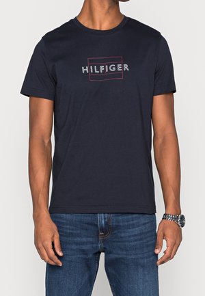 Homme portant un t-shirt bleu marine foncé avec le texte blanc ""HILFIGER"" encadré d'un fin rectangle rouge, associé à un jean bleu et une montre-bracelet en métal.