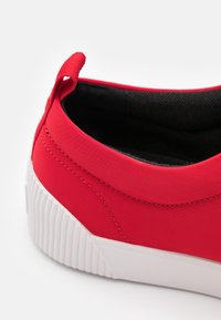 Rote Sportschuhe aus glattem synthetischem Material, mit einer abgerundeten Spitze, einer strukturierten weißen Gummisohle und einer Lasche an der Ferse.