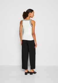 Witte mouwloze top met zwarte rand, gecombineerd met losse zwarte broek en zwarte hoge muiltjes. Model gezien van achteren.
