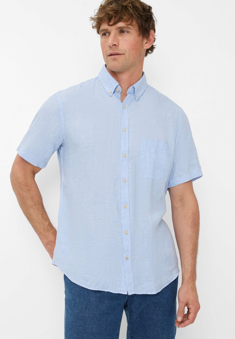 Camicia azzurro chiaro a maniche corte realizzata in tessuto testurizzato. Presenta un colletto con bottoni, una tasca sul petto e un orlo curvo.