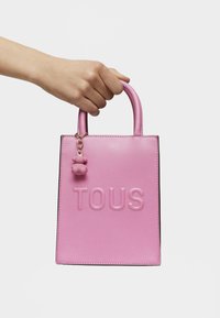 Sac fourre-tout en cuir rose avec logo "TOUS" embossé, poignées courtes et un charme en forme d'ours attaché. Texture lisse et forme structurée.