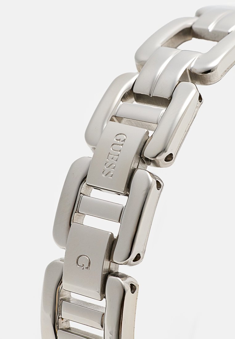Bracelet de montre en métal avec des maillons rectangulaires, finition argentée polie et logo "GUESS" gravé sur un maillon. Design lisse et moderne, texture uniforme.