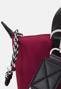 Malá kabelka s tělovým růžovým nylonovým tělem, černými koženými akcenty a pleteným černo-bílým páskem. Má stříbrné zapínání na zip.