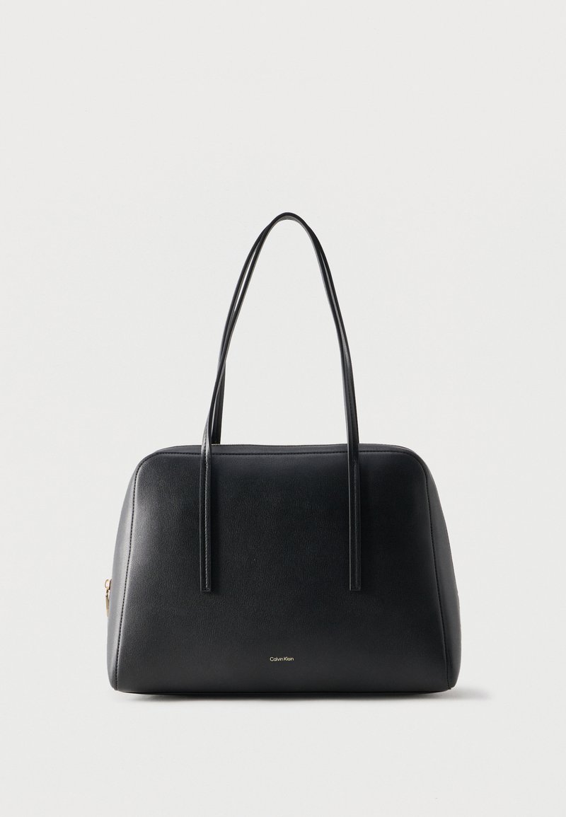 Μαύρη δερμάτινη τσάντα χειρός με δύο μακριές λουριά ώμου και μικρό λογότυπο Calvin Klein στο μπροστινό μέρος, απλό φωτεινό φόντο.