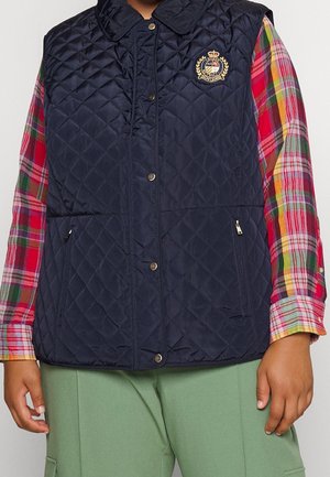 Personne portant un gilet matelassé bleu marine avec un emblème doré par-dessus une chemise à carreaux multicolores et un pantalon vert, visible de la poitrine jusqu'à mi-cuisse.
