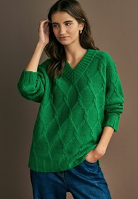 Maglione verde a maglia con un motivo a rombi, scollo a V e maniche oversize, abbinato a jeans blu, che mette in mostra il tessuto testurizzato.
