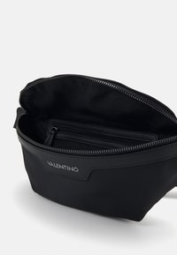 Musta nailonilaukku, jossa on vetoketjusulku, vuorattu sisäosa ja sisätasku. Brändäys näkyvillä edessä.