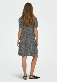 Schwarz Kleid mit weißem Blattmuster, kurzen Ärmeln und ausgestelltem Rock. Flache schwarze Schuhe vervollständigen das Outfit. Von hinten gesehen.
