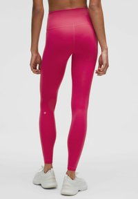 High-waisted, fitted leggings in bright pink, made of smooth, stretchy fabric. Notable logo detail near the ankle, paired with white athletic shoes.