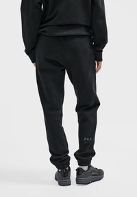 Schwarze Jogginghosen mit elastischen Bündchen, die ein dezentes "F.U.T." Logo-Detail aufweisen. Weicher Stoff mit lockerer Passform, kombiniert mit robusten schwarzen Turnschuhen.