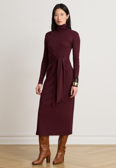Femme portant une robe bordeaux à col roulé manches longues avec une ceinture à la taille et des bottes marron à talons montant jusqu'aux genoux, debout sur un sol en bois.