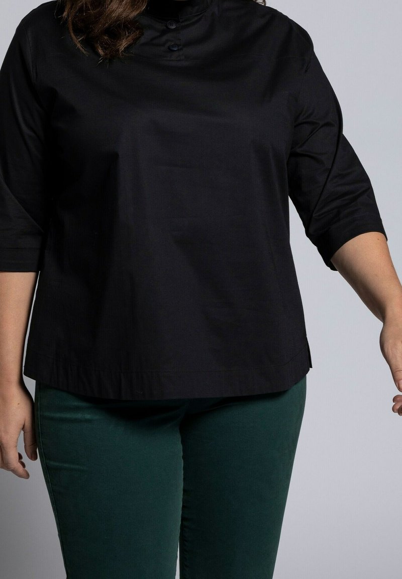 Personne portant un haut noir à manches trois-quarts et un pantalon vert foncé, debout devant un fond clair uni.