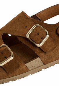 Hnědé semišové sandály se dvěma obdélníkovými kovovými přezkami. Otevřený design s výřezy a texturovanou béžovou podrážkou. Neformální a ergonomický styl.