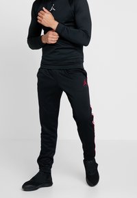 Completo sportivo nero composto da una maglia aderente a maniche lunghe e pantaloni abbinati; dettagli rossi lungo i lati e un logo sulla gamba sinistra.