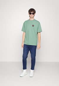 T-shirt de algodão verde com um pequeno logotipo quadrado no peito, combinado com jeans azul escuro e ténis brancos, usado por um modelo masculino.