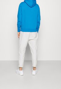 Persona vestida con una sudadera azul brillante, pantalones color crema enrollados en los tobillos y zapatillas blancas, de pie frente a una pared gris claro lisa.