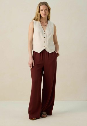 Gilet en lin crème avec cinq boutons, associé à un pantalon bordeaux à jambes larges. L'ensemble présente une coupe décontractée et des détails de design minimalistes.