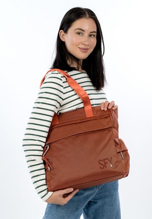 Frau trägt langärmliges gestreiftes Shirt und Jeans, hält eine große orangefarbene, strukturierte Tragetasche mit doppelten Griffen vor weißem Hintergrund.