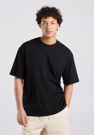 Jonge man met krullend donker haar draagt een loszittend zwart T-shirt en lichtbeige broek, staand tegen een effen grijze achtergrond.