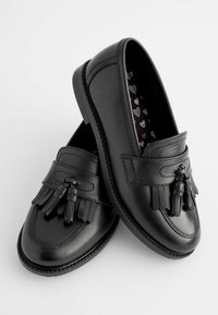 Des mocassins en cuir noir présentent un design de glands sur le dessus, une surface lisse et une semelle texturée. L'intérieur arbore un motif en forme de cœur.