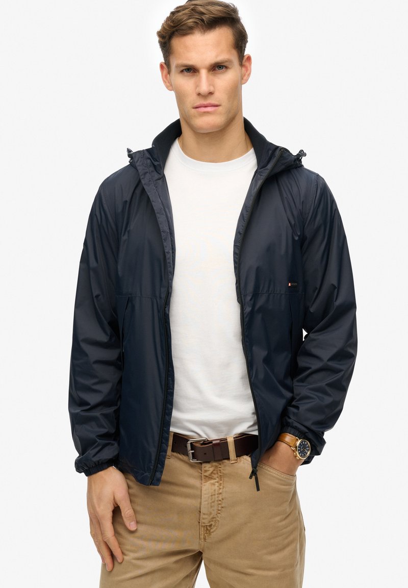 Navy windbreaker jacket with a hood, zip front, elastic cuffs, and two side pockets, worn over a white t-shirt and tan pants.