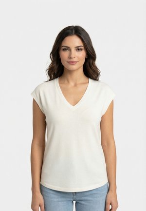 Mujer con cabello oscuro y ondulado que lleva una blusa blanca sin mangas con cuello en V y vaqueros azul claro, de pie contra un fondo blanco liso.