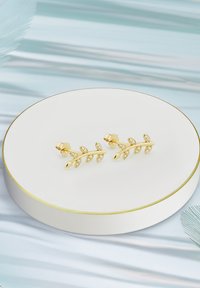 Örhängen med guldbladstema och klara stenar, uppvisade på en rund vit pedestal med guldkanter.