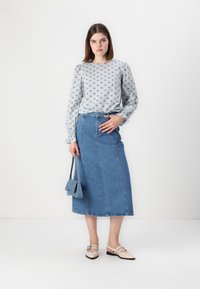 Blusa de manga longa azul claro com padrão, saia de ganga azul de cintura alta, sapatos bege com biqueira preta e uma pequena mala azul.