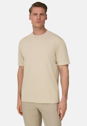 Mies pukeutuneena beigeihin lyhythihaiseen pyöreäkauluksiseen paitaan ja samanvärisiin housuihin, seisomassa yksinkertaisen vaaleanharmaan taustan edessä.