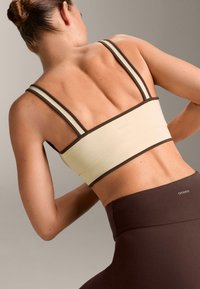 Vaaleabeige urheilurintaliivit, joissa on ruskeat yksityiskohdat, ohuet olkaimet ja sileä, joustava materiaali. Logo näkyy takana. Tummanruskeat legginsit.