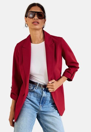 Kvinde iført oversized bordeaux blazer, hvid top, lyseblå jeans, mørkt bælte og store firkantede solbriller med et dugget glas.