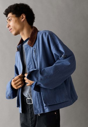Jeune homme aux cheveux bouclés portant une veste bleue avec un col marron sur une chemise rayée, ajustant la fermeture éclair de la veste sur un fond gris.