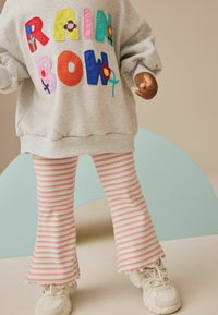 Grauer Sweatshirt mit gestickten, mehrfarbigen "RAINBOW"-Buchstaben und floralem Detail, kombiniert mit gestreiften, rosa-weißen Schlaghosen und weißen Turnschuhen.