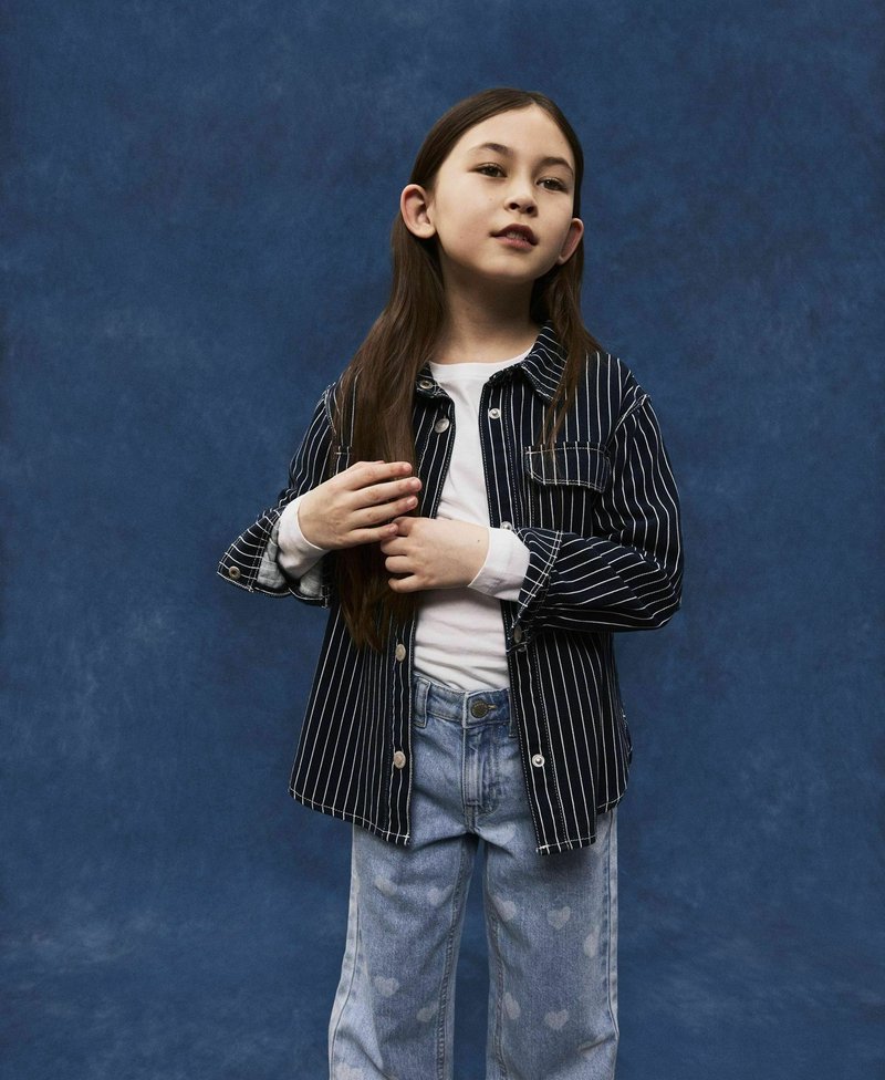 Chaqueta azul rayada de botones, camisa blanca de manga larga y jeans de lavado claro con patrones de corazones. El modelo se encuentra frente a un fondo azul.
