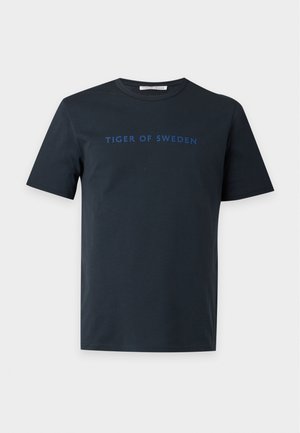 Mann trägt ein dunkelblaues "Tiger of Sweden" T-Shirt, dunkelbraune Hose und zweifarbige Loafers, steht vor einem einfarbigen hellen Hintergrund.
