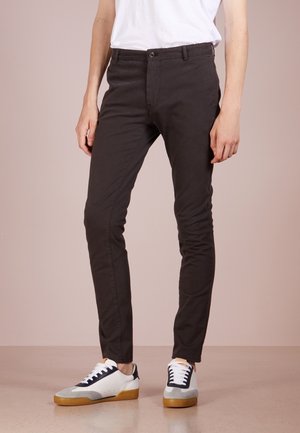 Person iført mørkebrune, tætsiddende bukser, hvid t-shirt og hvide sneakers med sorte detaljer, stående på en glat beige gulv.