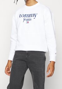 Sudadera blanca con logo azul de "Tommy Jeans", puños acanalados y cuello redondeado. Lleva unos pantalones oscuros gris. Diseño sencillo y casual.