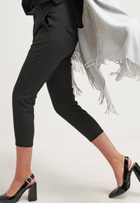 Černé kalhoty na míru s zkráceným lemem, doplněné lesklými černými lodičkami na podpatku. Světle šedý šál s třásněmi se skládá z jednoho ramene.