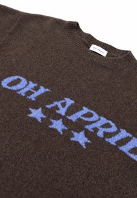 Καφέ πλεκτό πουλόβερ με γαλάζιο κείμενο "OH APRIL" και τρία μπλε αστέρια κάτω από το κείμενο, που δείχνει ριμπ πλεκτό στρογγυλό γιακά και ετικέτα.