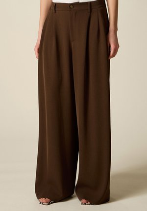 Pantalon large marron avec des plis et une fermeture à boutons, porté avec des talons ouverts, visible de la taille aux pieds sur un fond clair.