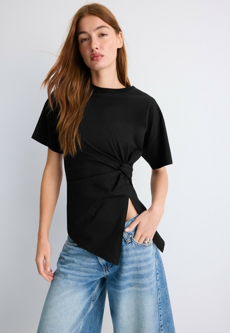 Jeune femme aux longs cheveux bruns porte un haut noir à manches courtes avec un nœud sur le côté et un jean large bleu clair, debout devant un fond uni.