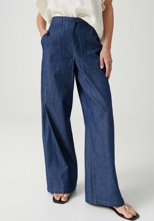 Weit geschnittene dunkelblaue Jeans mit aufgesetzten Fronttaschen, getragen zu weißem Top und schwarzen Zehensandalen.