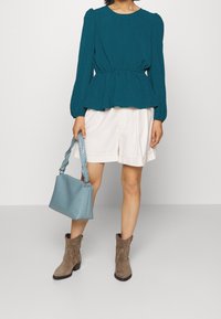 Blusa lunga teal a maniche lunghe con vita a peplum, abbinata a pantaloni beige. Borsa in tessuto azzurro chiaro con tracolla intrecciata e stivaletti marroni alla caviglia.