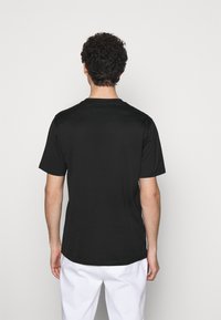 Mand med krøllet hår iført en ensfarvet sort kortærmet t-shirt og hvide bukser, stående mod en lys grå baggrund, vendt væk.